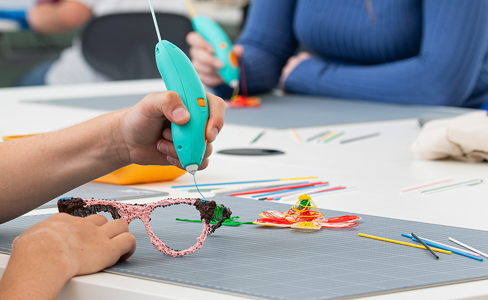 Kind sitzt an Tisch und “druckt” mit dem 3Doodler ein  Modell, andere Materialien liegen im Hintergrund